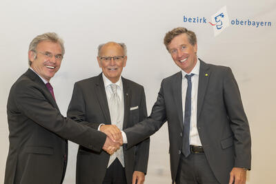 Drei Männer in Anzug geben sich die Hand. Im hinergrund das Logo des Bezirks Oberbayern.