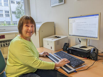 Eine Frau an ihrem Büroarbeitsplatz mit Computer, Monitor, Drucker, Tastatur sowie einer Brailletastatur davor.