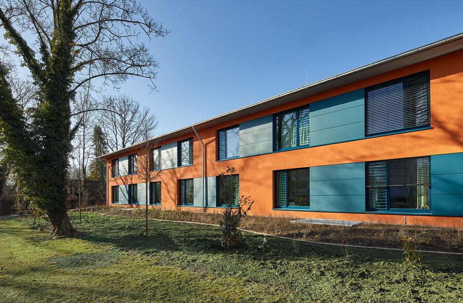 Auf einer grünen Grasfläche ist rechts ein zweistöckiges Gebäude zu sehen mit einer orangefarbenen Hausfassade. Die Fensterrahmen sind türkis.