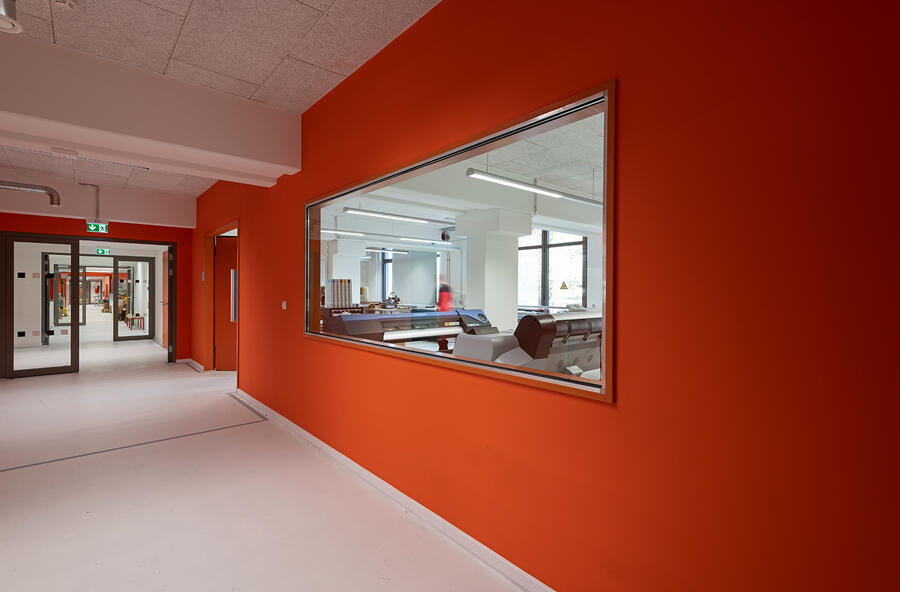 Links im Bild befindet sich ein langer Flur mit mehreren Glastüren. Die Wände sind in einem strahlenden Orange gestrichen. Rechts ist ein großes Glasfenster hinter dem sich ein weiterer Raum befindet.