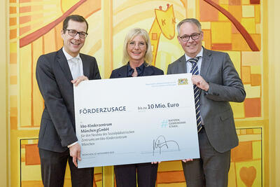 Drei Personen, zwei Männer und eine Frau halten ein Schild, das aussieht wie ein übergroßer Scheck, auf dem "Förderzusage" und "10 Mio. Euro" steht.