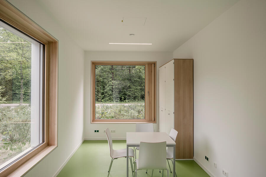 Ein heller Raum mit weißen Wänden und grünen Boden. In der Mitte steht ein weißer Tisch mit drei weißen Stühlen. Rechts im Eck steht ein schmaler Schrank. Links und hinten im Raum sind zwei große quadratische Fenster.