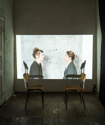Ein auf die Wand projeziertes Foto zeigt zwei Frauen, die sich gegenüber sitzen. Beide haben hellbraune Haare zu einem Dutt hoch gesteckt. Sie haben einen schwarzen Blazer an. Im Vordergrund des Raumes sieht man zwei Stühle, die sich gegenüber gestellt sind.