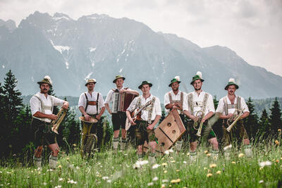 Sieben Musikanten in Tracht und mit ihren Instrumenten stehen in einer blühenden Sommerwiede, im Hintergrund erhebt sich ein Gebirge.