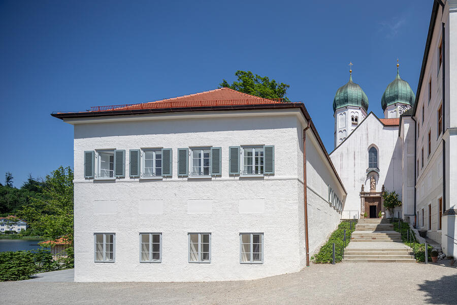 Links steht ein zweistöckiges Haus mit weißer Fassade. Die Fenster im oberen Stockwerk haben türkisfarbene Fensterläden. Rechts weiter hinten im Bild steht eine kleine Kirche mit zwei Türmen.