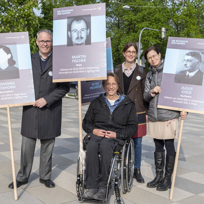 Ein Mann im Rollstuhl, zwei stehende Frauen und ein stehender Mann halten Schilder, auf denen ein Portrait, Namen und Informationen zu der auf dem Schid abgebnildeten Person vermerkt sind.