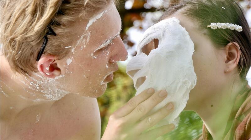 Junger Mann im Begriff eine Gipsmaske zu küssen, die er vor das Gesicht einer anderen Person hält. An seinem Gesicht sind noch die Gipsreste der Maske zu erkennen.