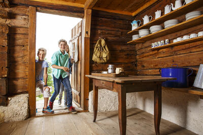 Drei Kinder betreten die Stube eines historischen Holzhauses mit Balken. Man sieht einen alten Tisch und Geschirr and den Wänden.