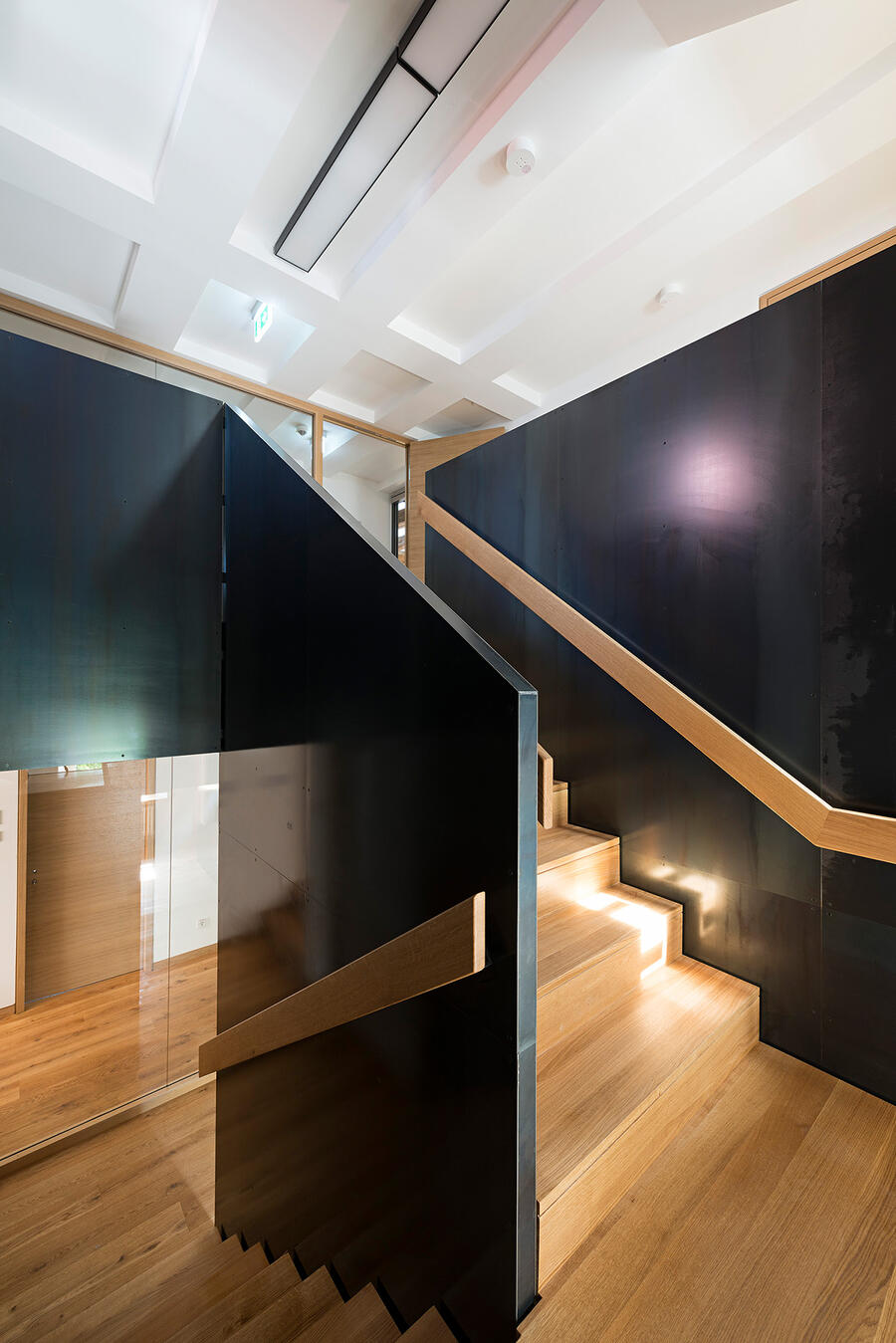 Ein Treppenaufgang mit schwarzen massiven Wänden. An den halbhohen Wänden sind helle Holzleisten als Geländer befestigt. Die Treppenstufen sind ebenfalls mit hellem Holz ausgelegt.