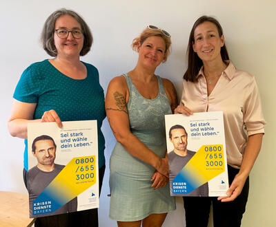 Drei Frauen mit zwei Plakaten. Aufschrift: Sei stark und wähle dein Leben: 0800/6553000. Krisendienste Bayern.
