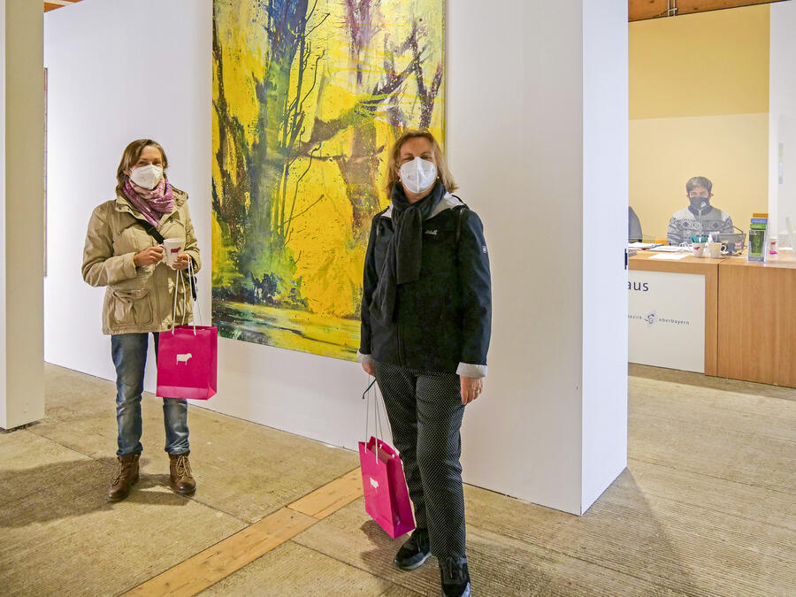 In einem Ausstellungsraum, an dessen Wänden großformatige Kunstwerke hängen, stehen zwei Besucherinnen mit FFP2-Gesichtsmasken. Im Hintergrund ist der Empfang zu sehen, an dem ein Mitarbeiter des Künstlerhauses hinter einer Plexiglasscheibe und ebenfalls mit FFP2-Maske zu sehen ist.