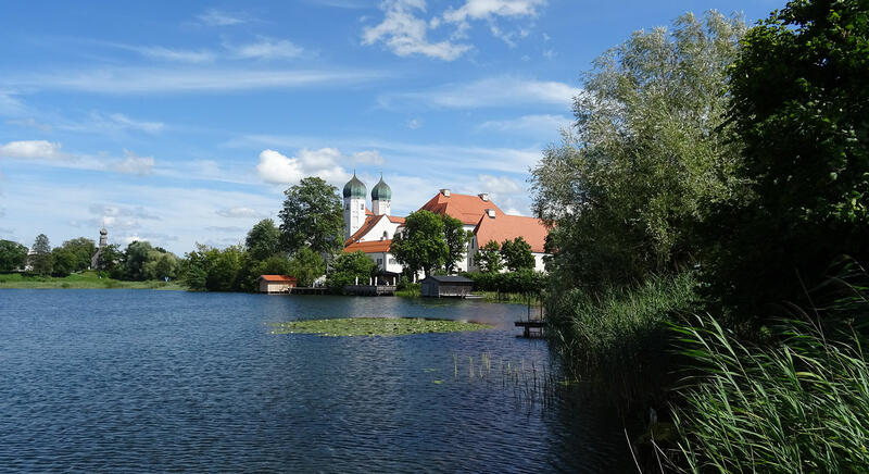 Landschaft mit See, Seerosen und Büschen im Vordergrund und den Kirchtürmen und historischen Gebäuden eines Klosters im Hintergrund
