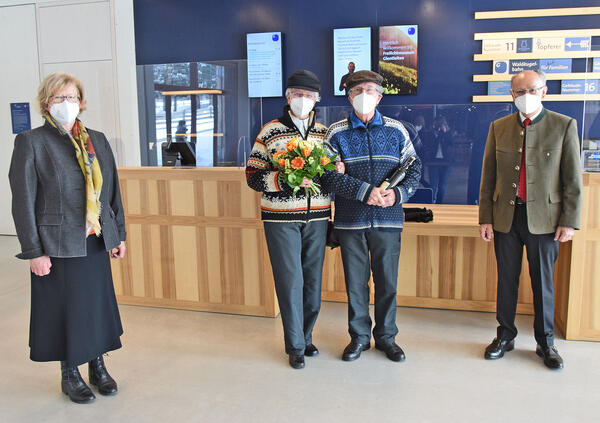 Ein älteres Paar mit Masken steht im Eingangsbereich des Museums Glentleiten. Die Frau trägt einen Blumenstrauß in der Hand. Rechts und links stehen Bezirkstagspräsident Josef Mederer und Museumsdirektorin Dr. Monika Kania-Schütz.
