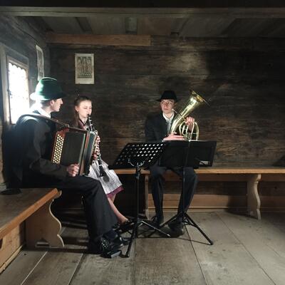 Ein Akkordeonspieler, eine Klarinettistin und ein Tubaspieler auf einer Eckbank in einer dunklen Bauernstube.