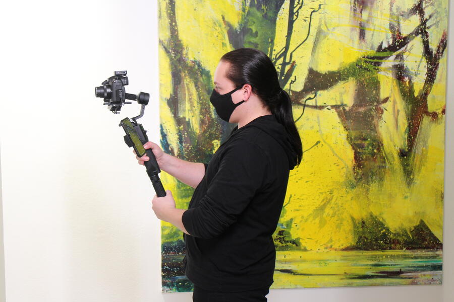 Ein junger Mann mit schwarzem Pullover und zurück gebundenen Haaren hält eine Filmkamera in der Hand.