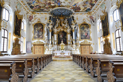 Blick in einen barocken Altarraum mit einem reichverzierten Altar und einer bemalten Decke. Im Vordergrund Holzbänke und ein Steinboden in Mosaikform.