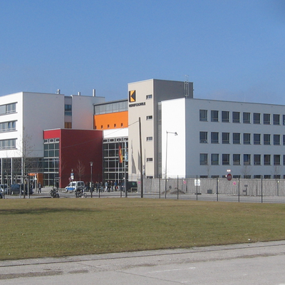 Außenansicht der Adolf-Kolping-Berufsschule in München:
Blick von einer Straße über einen Rasen auf einen Gebäude-Komplex aus Längs- und Querriegeln.   