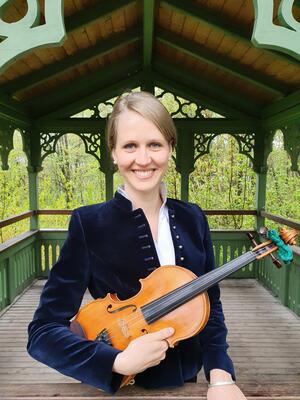 Lächelnde blonde Frau in blauer Samtjacke mit Geige unterm Arm steht in einem Pavillion-  