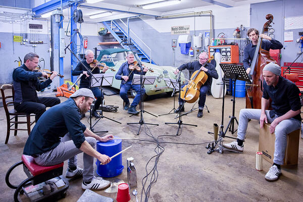 In einer Werkstatt sitzen sechs Männer in einem Halbkreis und spielen auf ihren Instrumenten. Im Hintergrund steht neben der Hebebühne ein Oldtimer. 