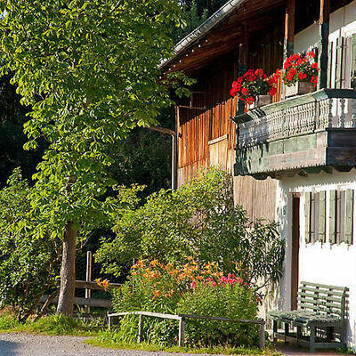 Foto eines Bauernhauses. Man sieht einen Teil der Fassade mit Holzbakon und Fensterläden in der Sonne sowie einen Baum und ein Blumenbeet vor dem Haus.