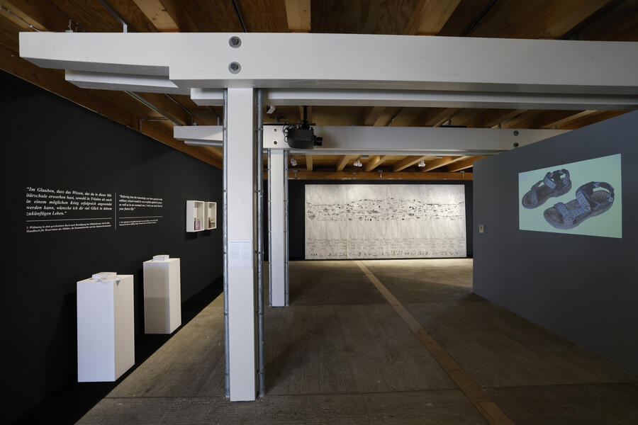 Blick in die Ausstellung mit Bildern und Text an den Wänden, einem großen Kunstwerk an der Wand, mehreren Stelen mit Kunstwerken