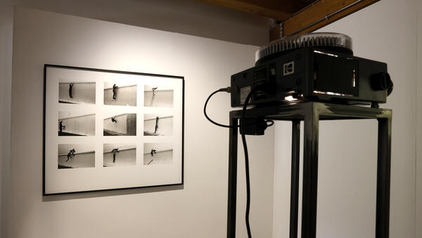 Ein Diaprojjektor beleuchtet einen Bilderrahmen, in dem sich neun kleine schwarz-weiss-Bilder befinden.