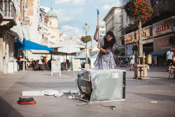 Kunstwerk: Eine Fotografie zeigt eine weibliche Person mit langen Haaren, die mit einem schweren Abbruchhammer auf eine liegende Waschmaschine einschlägt. Der Hintergrund legt nahe, dass sich diese Szene in einer Fußgängerzone abspielt, man sieht Passanten, Sonnenschirme, Tische und Stühle eines Kaffees.
