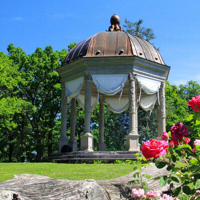 Sechseckiger Säulen-Pavilllon mit Bronzedach in einem Park. Im Vordergrund sind Heckenrosen, im Hintergrund Bäume zu sehen