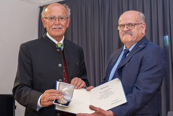 Zwei Männer in Anzügen mit Krawatte zeigen eine Urkunde und eine Medaille.