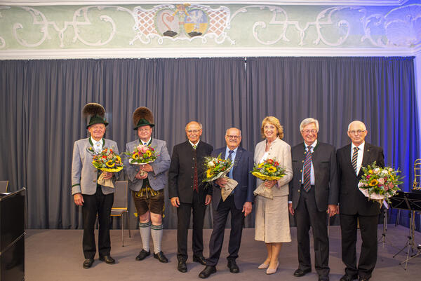 Eine Frau und sechs Männer vor stehen vor einem Bühnenvorhang.
Bis auf zwei Männer halten alle Personen einen Blumenstrauß in der Hand. Zwei Männer tragen Tracht.  