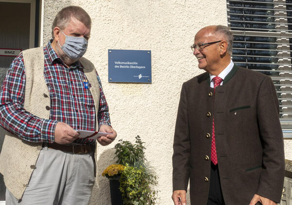 Zwei Männer stehen wor einem Gebäude, der linke trägt einen Mundschutz. 