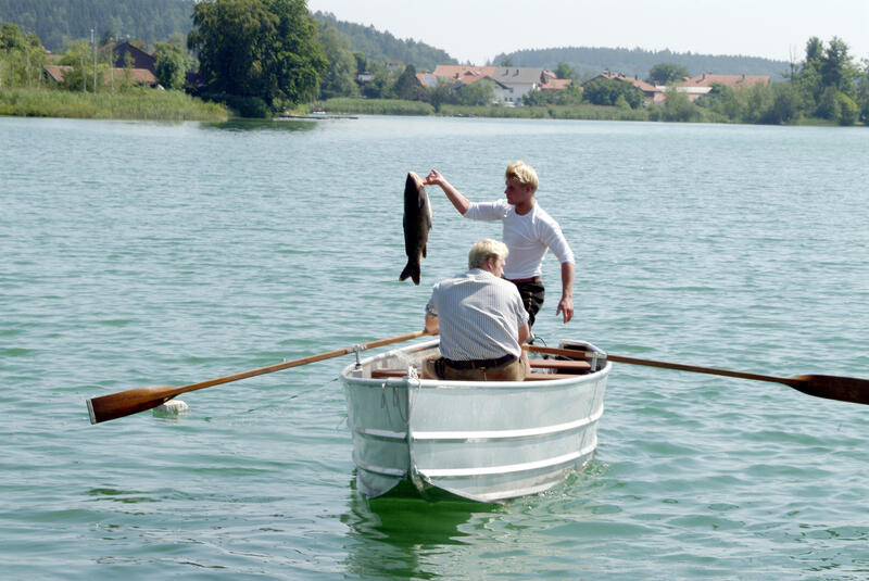 Foto eines Ruderbootes auf einem Gewässer. Auf dem Boot steht ein Mann, der einen großen Fisch ochhält. Mit dem Rücken zum betrachter sitzt ein zweiter Mann und rudert. Am Ufer sieht man Bäume und eine Ortschaft.